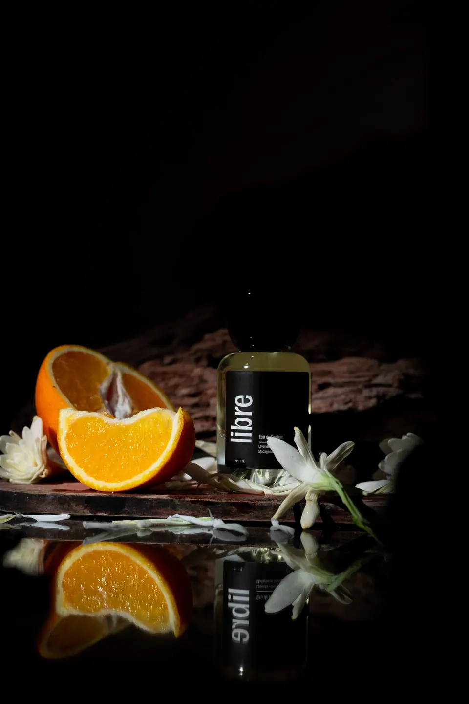 Foto produk parfum dengan irisan jeruk dan bunga di permukaan reflektif, foto oleh Capturels