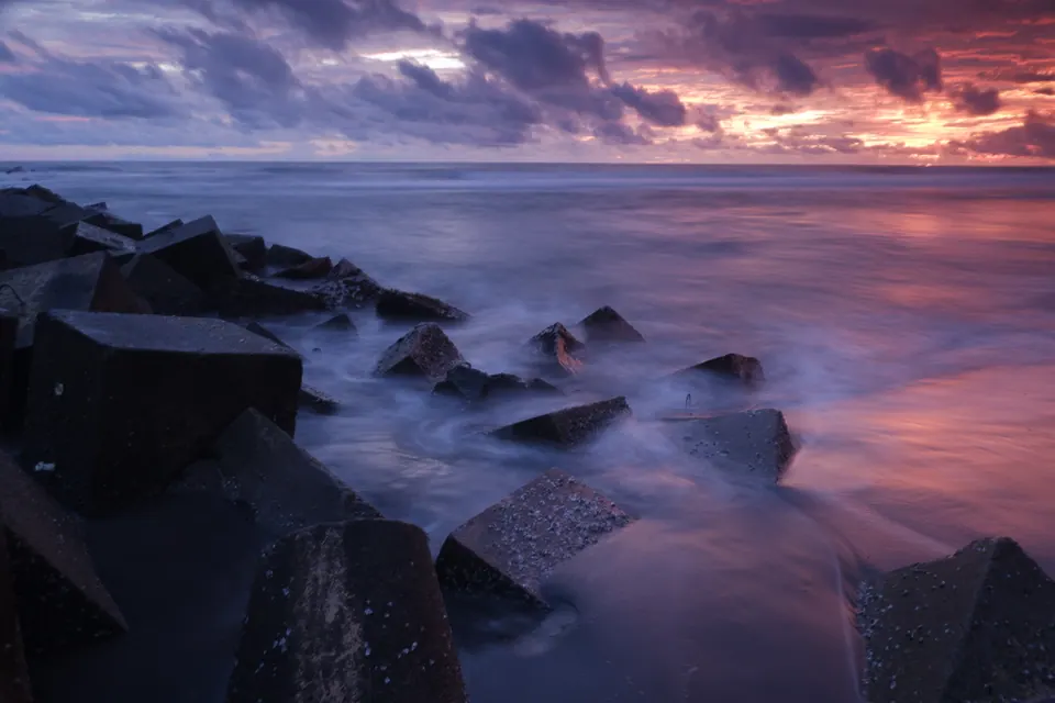 Foto lanskap pantai saat senja, ombak halus di antara batu pemecah gelombang, foto oleh Doeng