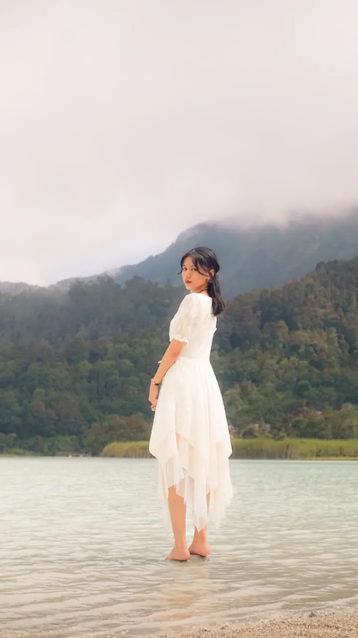 Portrait outdoor perempuan bergaun putih berdiri di tepi danau berkabut, foto oleh Hilenss