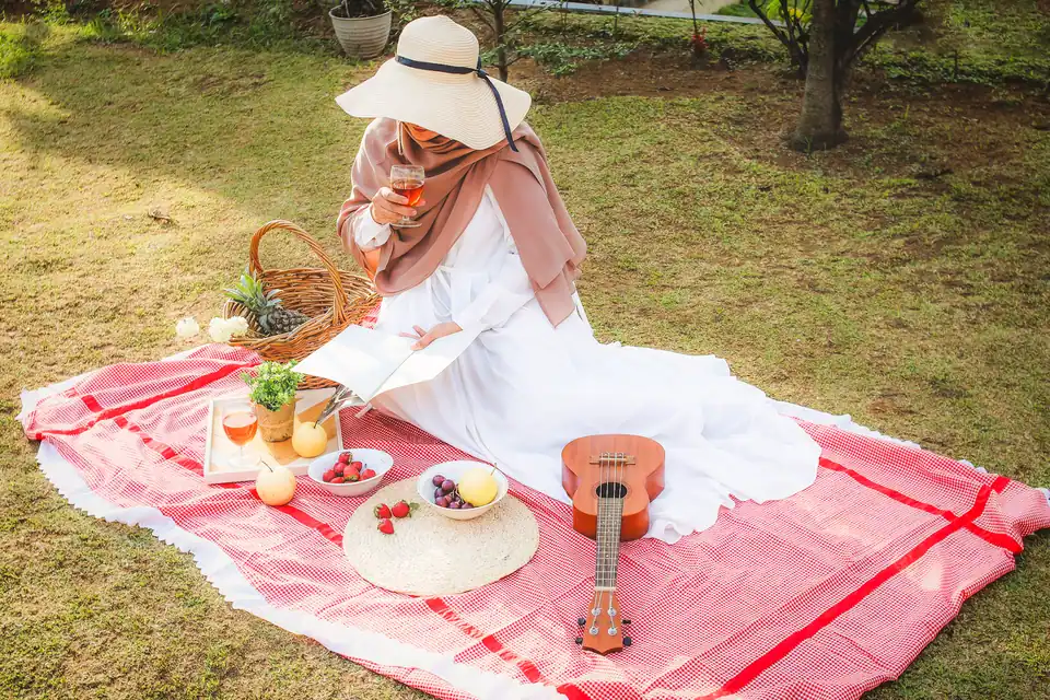 Foto konsep piknik: perempuan bertopi duduk di atas kain merah dengan buah dan ukulele, foto oleh Ihir