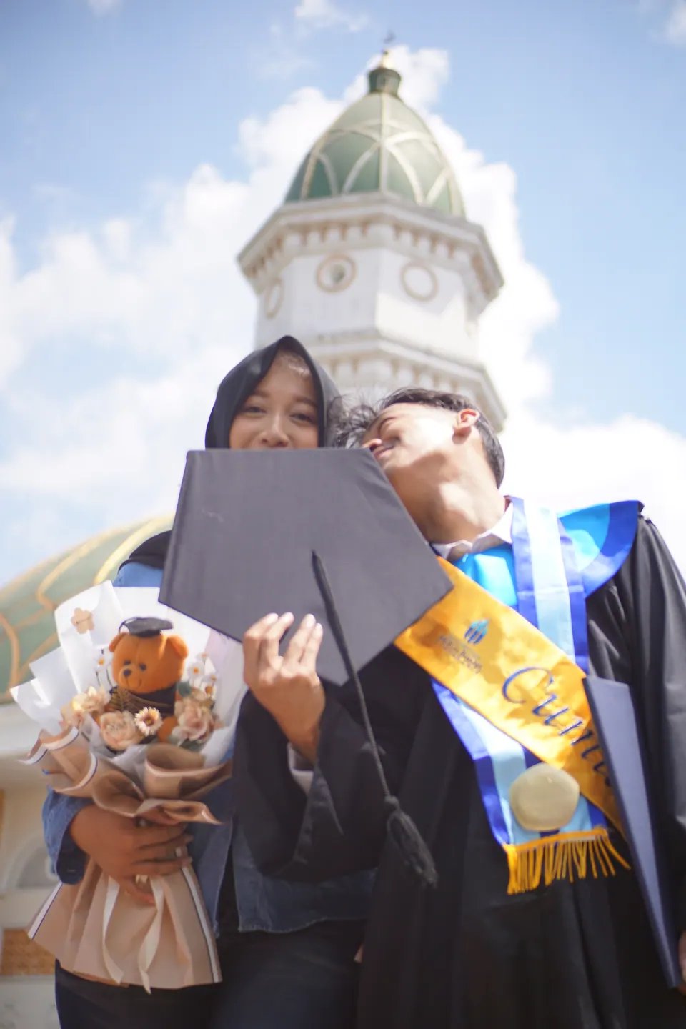 Foto wisuda: wisudawan bertoga berpose bersama ibunya di depan menara kampus, foto oleh Usman