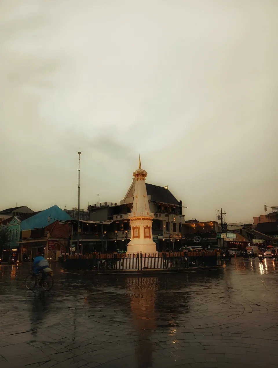 Foto jalanan kota setelah hujan dengan tugu dan pantulan di aspal basah, foto oleh Vier Studio