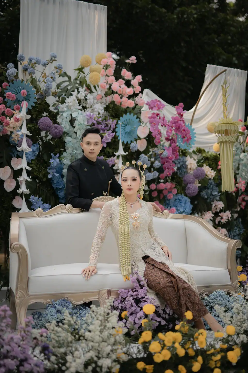 Foto pernikahan adat: pasangan duduk di sofa pelaminan penuh bunga, foto oleh Yusron