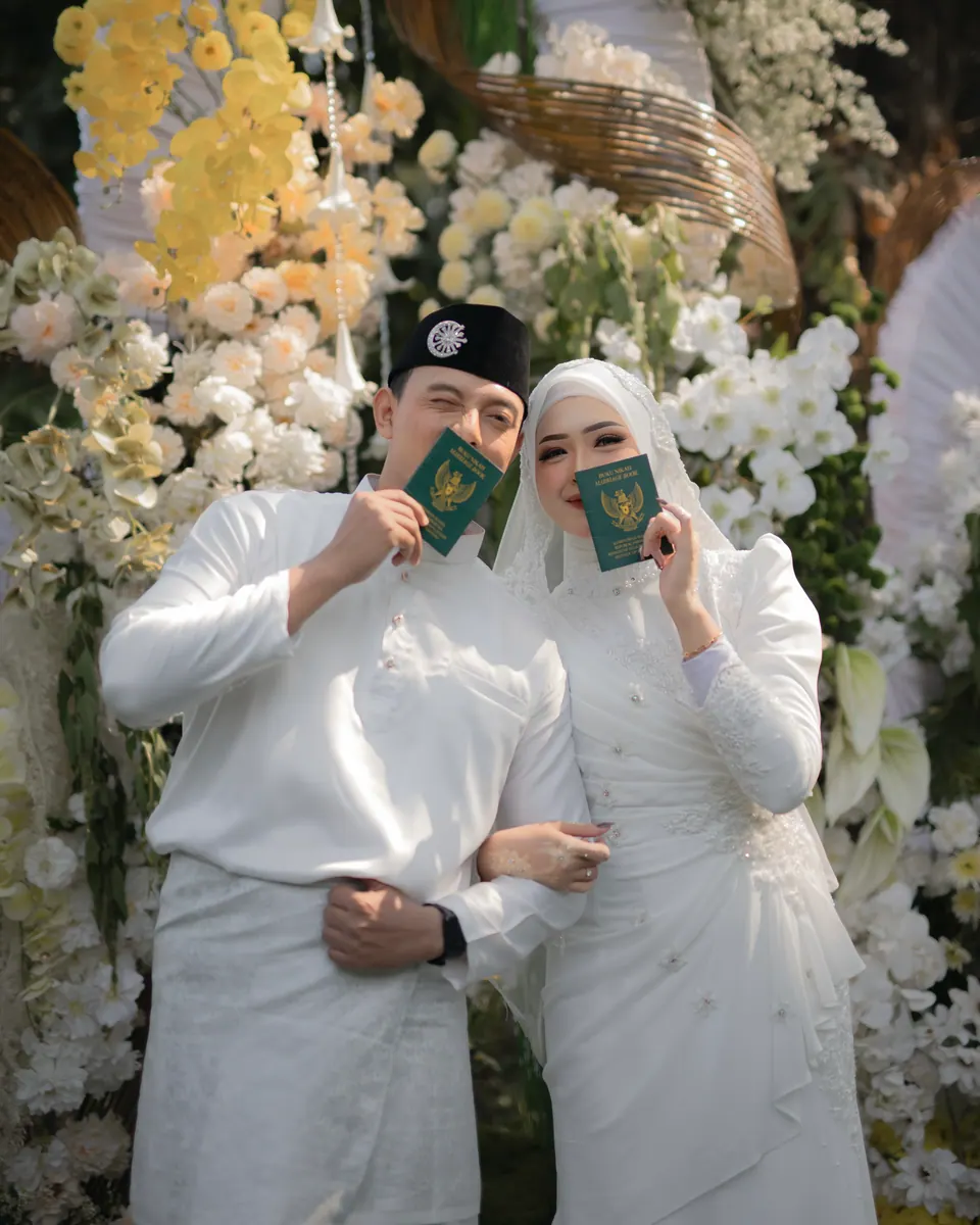 Foto pernikahan: pasangan pengantin berbusana putih memegang buku nikah, foto oleh Yusron